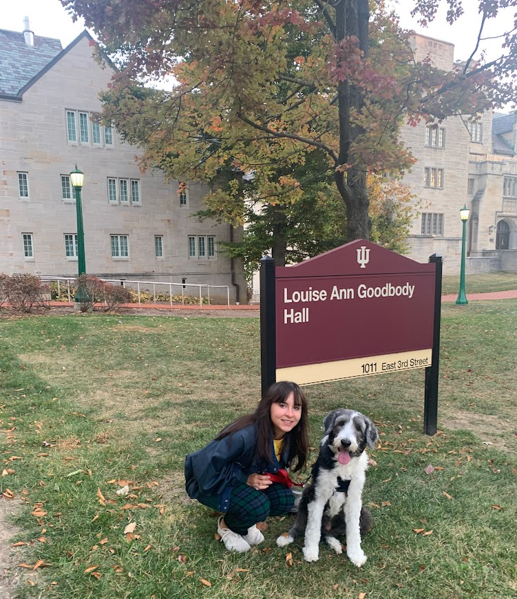A smiling young woman in a green plaid skirt and black jacket kneels beside a fluffy, black and white dog in front of the "Louise Ann Goodbody Hall" sign at 1011 East 3rd Street, with autumn-colored trees and a historic brick building in the background.
